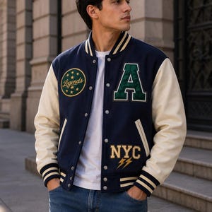 May include: A navy blue varsity jacket with cream-coloured sleeves, featuring a green letter A patch, a circular Legends patch with stars, and an NYC patch with lightning bolts. The jacket has a snap-button front, striped ribbed cuffs, and collar.