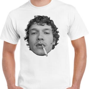 May include: White t-shirt with a black and white image of a person's face, showing curly hair and a cigarette. The graphic is centrally placed. The shirt has short sleeves and a crew neck.
