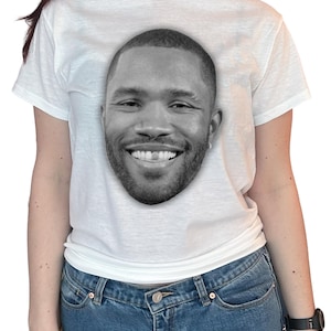 May include: White t-shirt featuring a black and white image of a smiling person's face. The graphic is centered on the front of the shirt. The shirt is made of a soft material and has short sleeves.