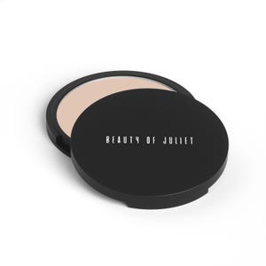 May include: A black compact with the text "BEAUTY OF JULIET" in white lettering. The compact is open, revealing a light beige powder. The lid is slightly ajar, casting a shadow on the white background.