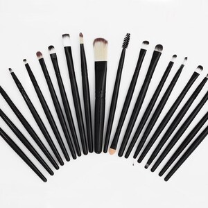 May include: A collection of makeup brushes arranged in a semi-circle on a white background. The brushes have black handles and various brush heads, including fluffy, angled, and pointed shapes. The bristles are in shades of white, brown, and beige.