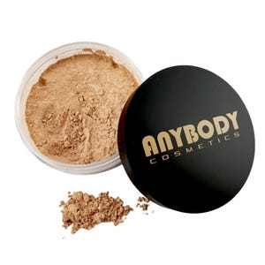 May include: An open jar of loose powder makeup with a black lid. The powder is a light brown shade, and a small pile of the powder sits next to the jar. The lid has the words "ANYBODY COSMETICS" printed in gold.