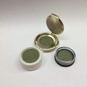 May include: Three eyeshadow compacts in varying styles. One is gold with a hinged lid, another is white, and the third is clear with a black base. All contain a matte olive green eyeshadow.