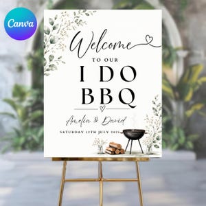May include: A white rectangular welcome sign featuring black text that reads Welcome to our I Do BBQ, Amelia & David, Saturday 12th July 2025. The design includes green watercolor foliage, a small black charcoal grill, and a stack of firewood.