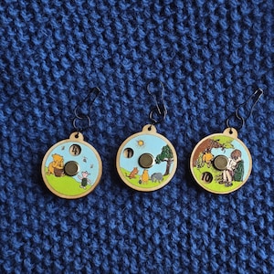 Könnte beinhalten: Drei Maschenmarkierer aus Holz mit Winnie-the-Pooh-Figuren. Jeder Marker hat eine andere Szene mit einer Nummer aus der Geschichte. Die Marker haben schwarze Metallverschlüsse und sind vor einem blauen Strickhintergrund platziert.