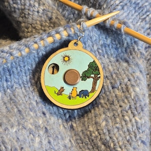 May include: A round wooden stitch marker with a Winnie the Pooh scene, including a sun, tree, and characters. It's attached to knitting needles and blue wool, with the number 1 showing.
