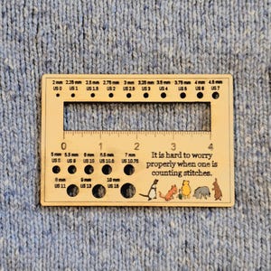 May include: A wooden knitting gauge with various hole sizes and millimeter measurements. The gauge includes the text "It is hard to worry properly when one is counting stitches" and small animal illustrations. The gauge is on a blue knitted background.