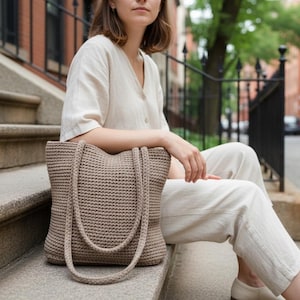 May include: A beige crochet tote bag with long handles. The bag is made of a thick, textured yarn and has a simple, functional design. The bag is sitting on a set of concrete steps. The bag is a neutral color.