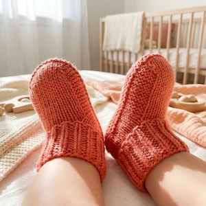 May include: A pair of coral-coloured knitted socks. The socks are thick and warm, with a ribbed cuff. The image is taken from a low angle, showing the socks on a bed with a cot in the background. The socks appear to be handmade.