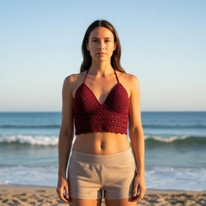 May include: A burgundy halter-neck crochet crop top with a V-neck design. The top is paired with beige shorts. The background features a beach scene with the ocean and sky.