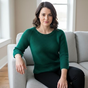 May include: A green knit sweater with a crew neck and long sleeves. The sweater is worn with black pants. The person is sitting on a gray couch in a well-lit room. The sweater is a solid color.