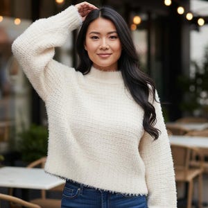 May include: A woman wearing a cream-colored, textured sweater with a mock neck and long sleeves. The sweater is paired with blue jeans. The background includes outdoor seating and greenery.