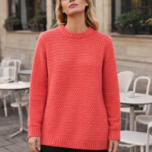 May include: A coral-colored sweater with a textured knit pattern. The long-sleeved sweater has a crew neck and ribbed cuffs and hem. The model is wearing the sweater in an outdoor setting with tables and chairs in the background.