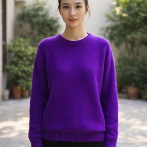 May include: A vibrant purple sweater with a crew neck. The sweater is made of a soft, textured material. The model is wearing a black bottom. The background features green foliage and potted plants.