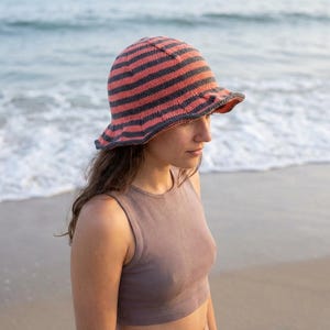 May include: A striped cloche hat in coral and navy blue, with a small brim. The hat is worn by a person on a beach, with the ocean in the background. The hat is a stylish accessory.