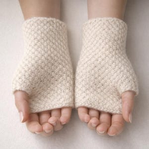 May include: A pair of off-white, fingerless gloves made of a textured knit fabric. The gloves cover the hand up to the knuckles, leaving the fingers free. The gloves are shown against a neutral background.