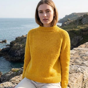 May include: A woman wearing a mustard yellow knit sweater with a mock neck. The sweater has long sleeves and a ribbed hem. The background features a rocky coastline and the ocean.
