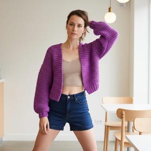 Puede incluir: Una mujer con una rebeca de punto morado con mangas abullonadas y pantalones cortos vaqueros azul oscuro. La rebeca está abierta, revelando un top beige. El fondo incluye una zona de cocina con estanterías de madera y una mesa blanca.