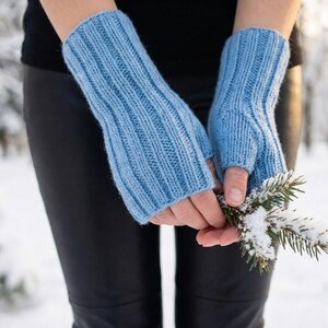 May include: Light blue knitted fingerless gloves with a ribbed design. The gloves are made of a soft, warm material, perfect for cold weather. The gloves are shown with a snowy background and a sprig of evergreen.