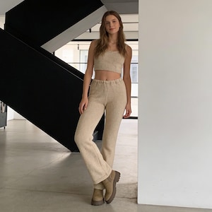 May include: A beige two-piece outfit consisting of a crop top and matching pants. The outfit is made of a textured fabric. The model is wearing brown boots. The background includes a black staircase and a white wall.