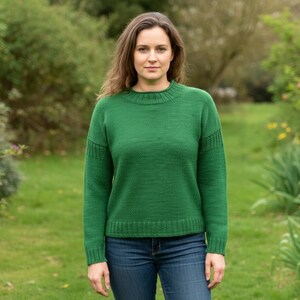 May include: A woman wearing a green crew neck sweater and blue jeans stands in a garden. The sweater has ribbed detailing on the cuffs and neckline. The background features green foliage and a grassy area.