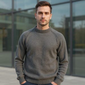 May include: A gray crew neck sweater with raglan sleeves. The sweater is made of a knitted material and has a ribbed collar, cuffs, and hem. The person is wearing the sweater and has their hands in their pockets.