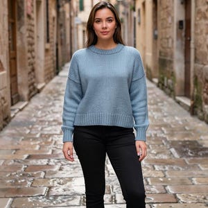 May include: A light blue sweater with ribbed detailing is paired with black pants. The sweater has a crew neck and long sleeves. The model is standing on a cobblestone street.