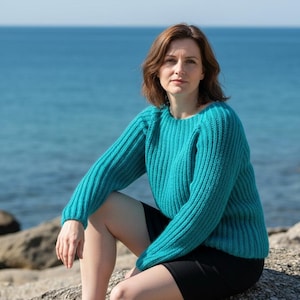 May include: A teal ribbed knit sweater with long sleeves. The sweater has a crew neck and is worn with a black skirt. The background features a blue ocean and rocks.