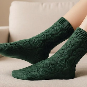 May include: A pair of dark green knit socks with a cable-knit pattern. The socks are thick and appear warm, with a ribbed cuff at the top. The socks are on a light-colored couch.