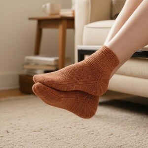 May include: A pair of knitted socks in a warm, earthy brown color. The socks are visible on the feet of a person, with the legs resting on a beige sofa. The socks have a textured knit pattern.