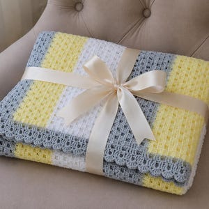 Puede incluir: Una manta de bebé hecha a mano, doblada, con una combinación de colores amarillo, blanco y gris. La manta está atada con una cinta color crema. La manta tiene un borde festoneado. Mide aproximadamente 90 cm x 90 cm.