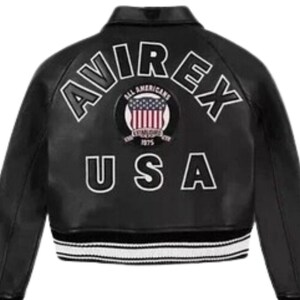 Puede incluir: Chaqueta de cuero negra con letras blancas que dicen "AVIREX" y "USA". La chaqueta presenta un parche con la bandera estadounidense y las palabras "ALL AMERICANS" y "1975". La chaqueta tiene un dobladillo a rayas blancas y negras.