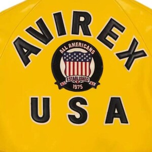 May include: A yellow jacket with black and white lettering and patches. The jacket displays the text "AVIREX" and "USA". A circular patch shows "ALL AMERICANS" and "ESTABLISHED 1975" with a US flag design.
