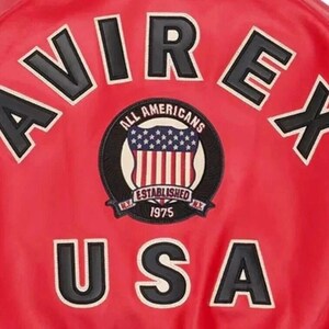 Puede incluir: Chaqueta roja con letras en blanco y negro y un parche circular. El parche dice "ALL AMERICANS ESTABLISHED 1975 N.Y." con un dise&ntilde;o de la bandera estadounidense. La palabra "USA" se muestra en letras grandes.