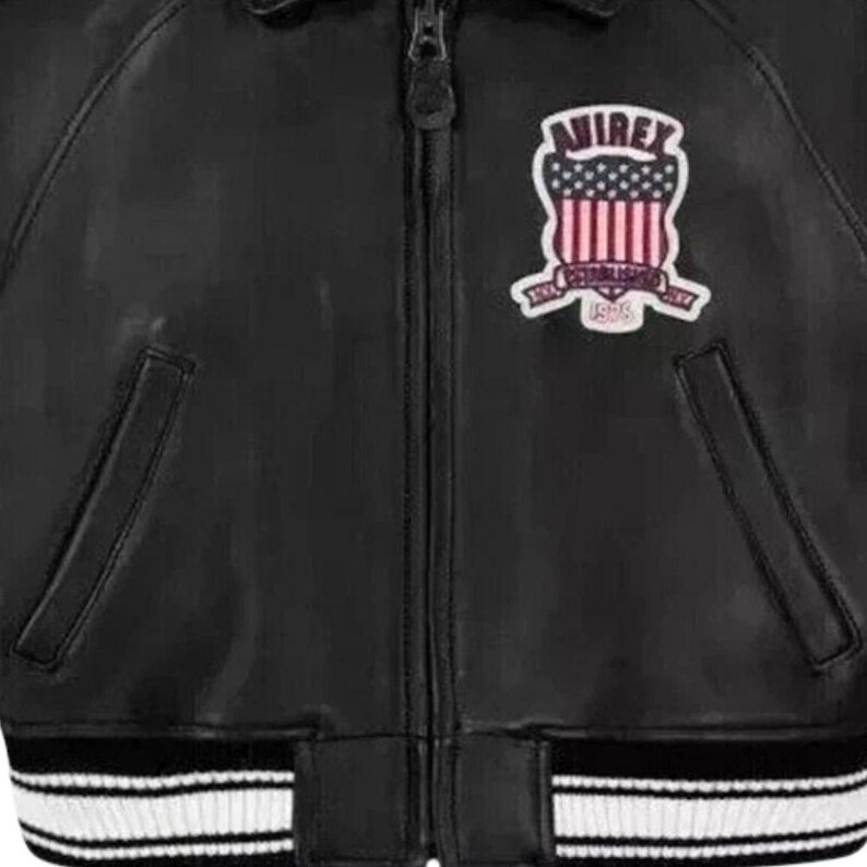 Puede incluir: Chaqueta de cuero negra con cierre de cremallera y dobladillo a rayas blancas y negras. La chaqueta presenta un parche en el pecho con el texto "AVIREX" sobre un dise&ntilde;o de rayas y estrellas rojas y blancas.