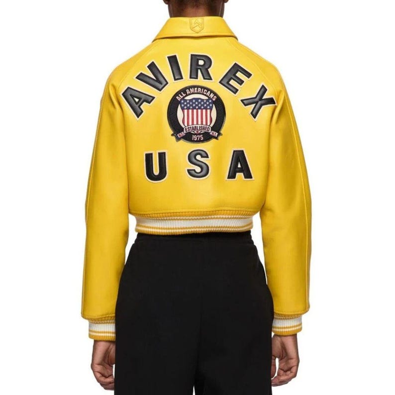 May include: A cropped yellow leather jacket with the word "AVIREX" arched across the top and "USA" at the bottom. The back of the jacket features a circular emblem with the words "ALL AMERICANS ESTABLISHED 1975". The jacket has white and yellow striped cuffs.