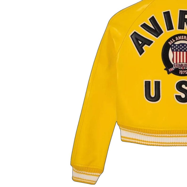 May include: Yellow varsity jacket with black lettering spelling out "AVIR" and "US". The jacket features a patch with the American flag and the words "ALL AMERICAN" and "ESTABLISHED 1975". The cuffs and hem are white and yellow striped.