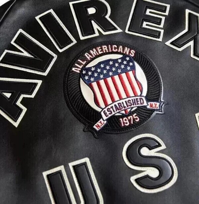Puede incluir: Chaqueta de cuero negra con letras blancas y negras que deletrean "AIREX" y "US". Un parche circular presenta la bandera estadounidense y las palabras "ALL AMERICANS ESTABLISHED 1975 N.Y."