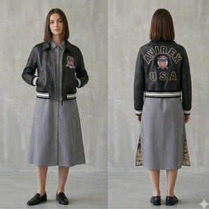 Puede incluir: Chaqueta de cuero negra con ribete a rayas blancas y negras y parche a juego. La chaqueta se lleva sobre un vestido a rayas. La parte trasera de la chaqueta tiene el texto "AVIREX USA" con un emblema patri&oacute;tico.