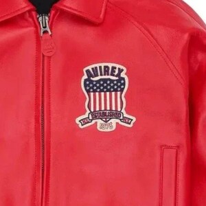 Puede incluir: Chaqueta de cuero roja brillante con cierre de cremallera. La chaqueta presenta un parche en el pecho con el texto "AVIREX" sobre un dise&ntilde;o de la bandera estadounidense y las palabras "ESTABLISHED 1975".