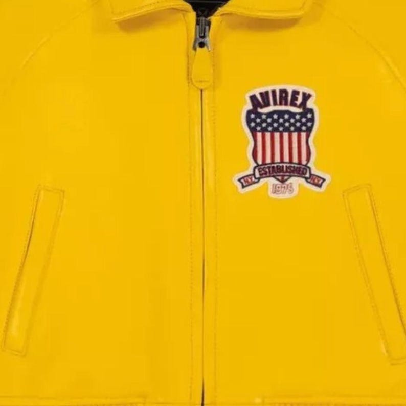 May include: A close-up of a bright yellow jacket with a zip. The jacket has a patch with the text "AVIREX" above a design of red and white stripes and blue stars. The patch also includes the text "ESTABLISHED 1975".