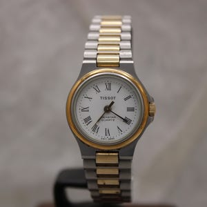 May include: A Tissot watch with a silver and gold-tone metal band and a white face. The watch has a gold-tone bezel and Roman numeral hour markers. The watch is a quartz model.