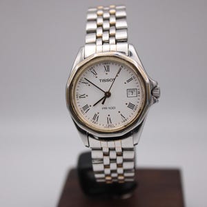 May include: A silver and gold Tissot PR 100 wristwatch with a white face, Roman numeral hour markers, and a date window at the 3 o'clock position. The watch has a stainless steel band and a gold-tone bezel. The watch is displayed on a dark brown stand.