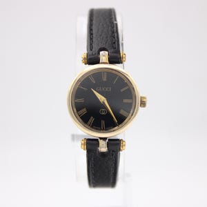 Könnte beinhalten: Eine goldfarbene Gucci-Armbanduhr mit schwarzem Zifferblatt und goldenen Zeigern. Die Uhr hat ein schwarzes Lederarmband. Das Zifferblatt zeigt das Gucci-Logo. Die Uhr hat ein rundes Zifferblatt und eine kleine Krone.