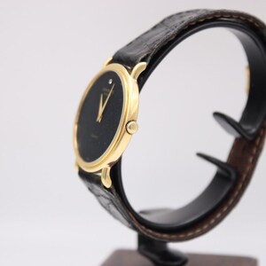 Angled side view of the Raymond Weil watch showing its slim gold case and black leather strap displayed on a watch stand.