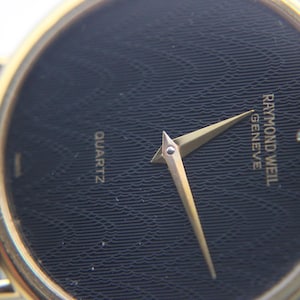 Macro shot of the black textured dial with a subtle wave pattern, gold tone hands, and the single diamond marker at 12 o'clock.
