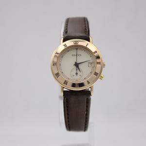 Op de afbeelding: Een goudkleurig Gucci-horloge met een crèmekleurige wijzerplaat, Romeinse cijfers en een datumweergave. Het horloge heeft een bruine leren band. De wijzerplaat heeft een diameter van ongeveer 3,2 cm.