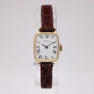 Puede incluir: Un reloj de pulsera Ingersoll vintage con una caja rectangular dorada y una esfera blanca con números romanos negros. El reloj tiene una correa de cuero marrón. La esfera del reloj dice "Ingersoll".