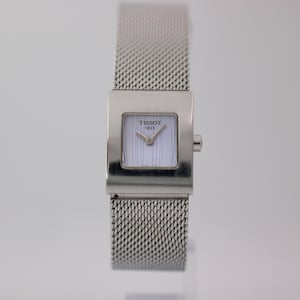 Op de afbeelding: Een zilverkleurig Tissot 1853 polshorloge met een vierkante kast en een fijne metalen mesh-band. De wijzerplaat is wit met dunne verticale lijnen, zilveren wijzers en het Tissot 1853-logo in zwarte letters in het midden bovenaan.