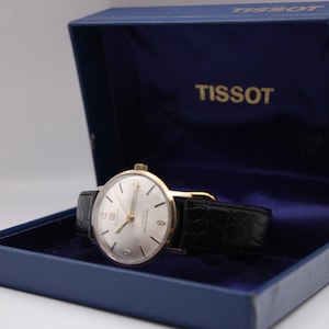 Może przedstawiać: Zegarek Tissot w stylu vintage ze srebrną tarczą i złotymi wskaz&oacute;wkami, umieszczony w niebieskim pudełku. Zegarek posiada czarny sk&oacute;rzany pasek. Otwarte pudełko eksponuje zegarek i nazwę marki "TISSOT".